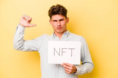Sarı arka planda izole edilmiş NFT levhası tutan genç beyaz adam gururlu ve kendinden emin hissediyor, örnek olarak.