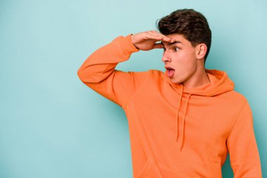 Kafkasyalı genç adam mavi arka planda izole edilmiş, uzaklara bakıyor ve elini alnına koyuyor..