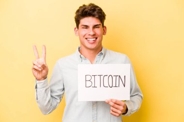 Sarı arka planda izole edilmiş bitcoin pankartı tutan beyaz bir adam neşeli ve tasasız parmakları olan bir barış sembolü gösteriyor..