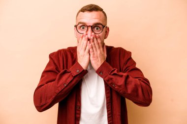 Beyaz tenli genç adam bej arka planda izole edilmiş şok olmuş, ağzı ellerle kaplanmış, yeni bir şey keşfetmeye hevesli..