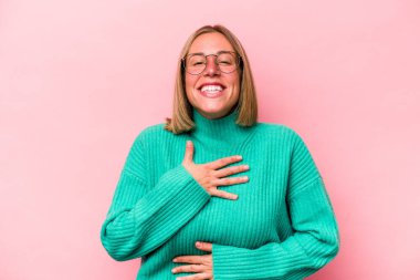 Pembe arka planda izole edilmiş genç beyaz kadın mutlu bir şekilde gülüyor ve ellerini karnının üstünde tutarak eğleniyor..