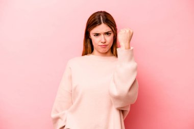 Pembe arka planda izole edilmiş genç beyaz kadın yumruğunu kameraya gösteriyor, agresif yüz ifadesi..