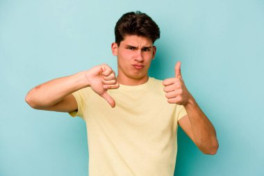 Kafkasyalı genç adam mavi arka planda izole edilmiş, baş parmağını kaldırıyor, zor seçim konsepti.