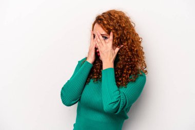 Beyaz arka planda izole edilmiş genç, beyaz bir kadın parmaklarının arasından korkmuş ve gergin bir şekilde göz kırpıyor..