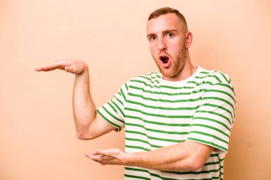 Beyazlı genç bir adam bej arka planda izole edilmiş. Elleri arasında bir kopya alanı tutarken şok olmuş ve hayrete düşmüş..