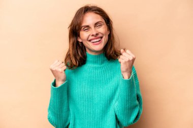 Beyaz tenli genç bir kadın bej arka planda izole edilmiş tasasız ve heyecanlı bir şekilde tezahürat yapıyor. Zafer konsepti.