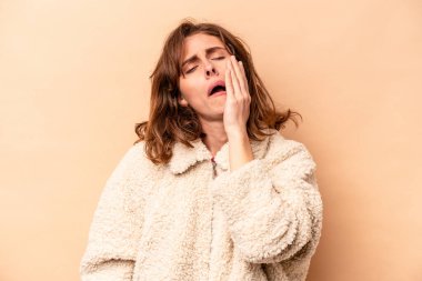 Beyaz tenli genç bir kadın bej arka planda izole edilmiş. Yorgun ve uykulu. Elini başının üstünde tutuyor..