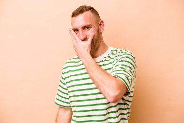 Beyazlı genç bir adam bej arka planda izole edilmiş. Kameraya parmaklarıyla göz kırpıyor, yüzünü kapatmaktan utanıyor..