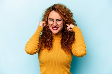 Kızıl saçlı genç bir kadın mavi arka planda izole edilmiş parmaklarıyla kulaklarını kapatıyor, gürültülü bir ortam yüzünden stresli ve çaresiz..