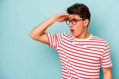 Kafkasyalı genç adam mavi arka planda izole edilmiş, uzaklara bakıyor ve elini alnına koyuyor..