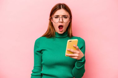 Elinde cep telefonu olan beyaz bir kadın pembe arka planda izole edilmiş çok kızgın ve agresif çığlıklar atıyor..