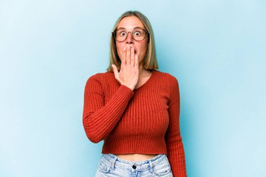 Mavi arka planda izole edilmiş genç beyaz kadın şok olmuş, ağzı ellerle kaplanmış, yeni bir şey keşfetmeye hevesli..