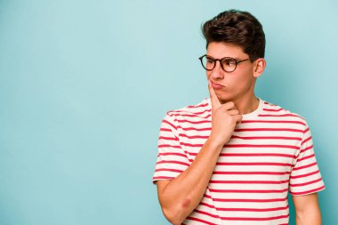Mavi arka planda izole edilmiş genç beyaz adam şüpheli ve şüpheci bir ifadeyle yana bakıyor..