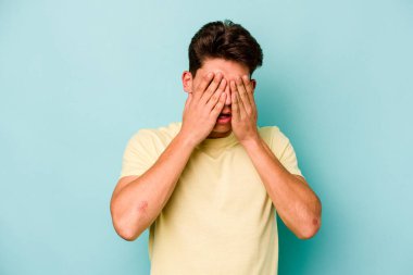 Mavi arka planda izole edilmiş genç beyaz adam elleriyle gözlerini kapatmaktan korkuyor..