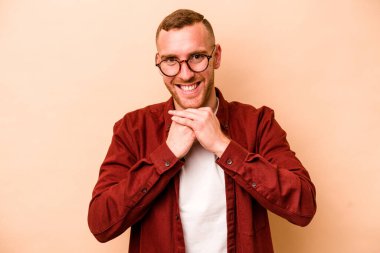 Beyaz tenli genç adam bej arka planda izole edilmiş ellerini çenesinin altında tutuyor, mutlu bir şekilde yana bakıyor..