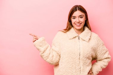 Pembe arka planda izole edilmiş genç beyaz kadın işaret parmağıyla işaret ederek gülümsüyor..