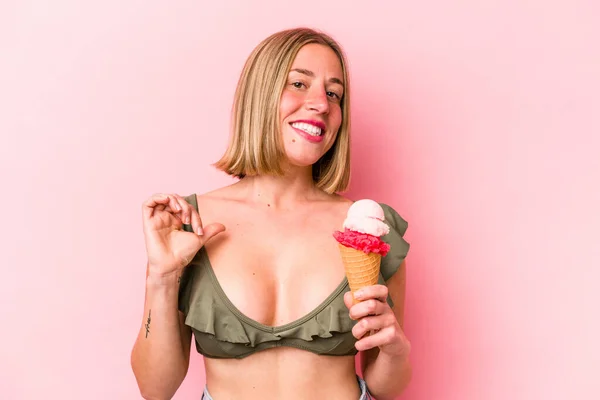 若い白人女性はビキニを着てピンクの背景に隔離されたアイスクリームを保持することは誇りと自信を感じています 次の例に従うこと — ストック写真