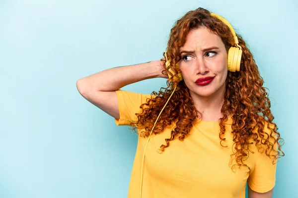 Young ginger woman listening to music isolated on blue background touching back of head, thinking and making a choice.
