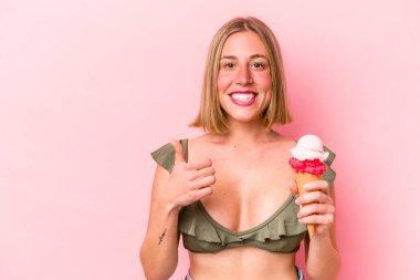 Beyaz bir kadın bikini giyiyor ve elinde pembe arka planda izole edilmiş bir dondurma tutuyor gülümsüyor ve başparmağını kaldırıyor.