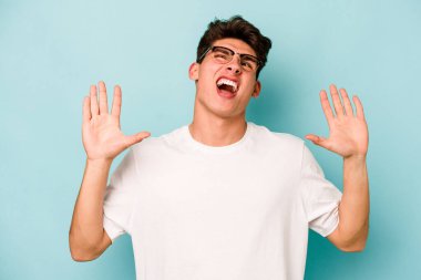 Kafkasyalı genç adam mavi arka planda izole edilmiş gökyüzüne haykırıyor, yukarı bakıyor, hayal kırıklığına uğramış..