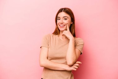 Pembe arka planda izole edilmiş genç beyaz kadın gülümsüyor ve kendinden emin, elleriyle çenesine dokunuyor..
