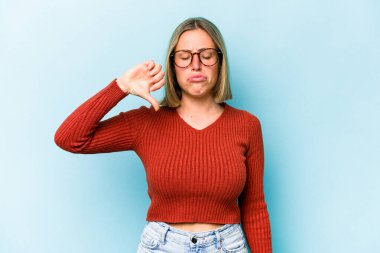 Mavi arka planda izole edilmiş genç beyaz kadın baş parmağını aşağı gösteriyor, hayal kırıklığı konsepti.