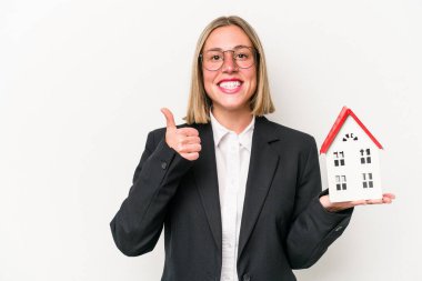 Beyaz arka planda izole edilmiş oyuncak evi tutan beyaz bir kadın gülümsüyor ve başparmağını kaldırıyor.