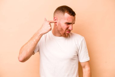 Beyazlı genç bir adam bej arka planda izole edilmiş kulakları elleriyle kapatıyor..