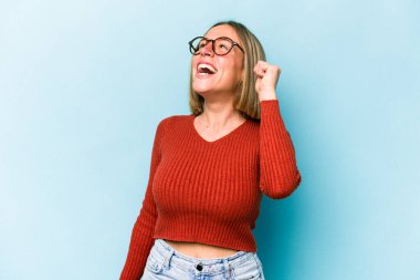 Mavi arka planda izole edilmiş genç beyaz kadın zaferi, tutkuyu ve coşkuyu kutluyor, mutlu bir ifade..