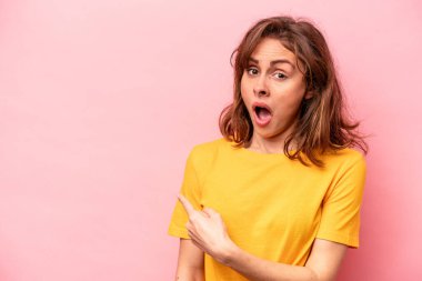 Pembe arka planda izole edilmiş genç beyaz kadın gülümsüyor ve boş alanda bir şeyler gösteriyor..