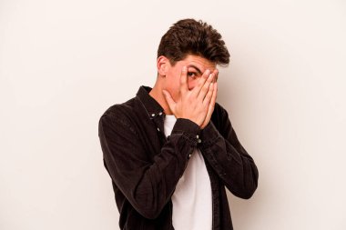 Beyaz arka planda izole edilmiş genç beyaz bir adam parmaklarının arasından korkmuş ve gergin bir şekilde göz kırpıyor..