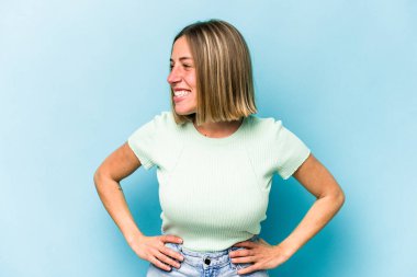 Mavi arka planda izole edilmiş genç beyaz kadın mutlu bir şekilde gülüyor ve ellerini karnının üstünde tutarak eğleniyor..