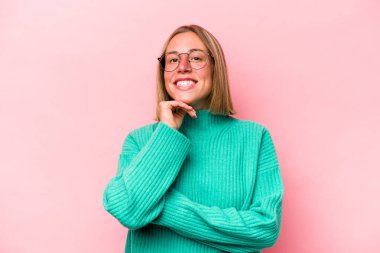 Pembe arka planda izole edilmiş genç beyaz kadın gülümsüyor ve kendinden emin, elleriyle çenesine dokunuyor..