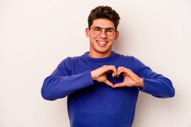 Beyaz arka planda izole edilmiş genç beyaz bir adam gülümsüyor ve elleriyle kalp şekli gösteriyor..