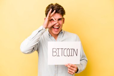 Sarı arka planda Bitcoin levhası tutan beyaz bir adam heyecanlı. Göze göz işareti yapıyor..
