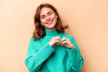 Beyaz tenli genç bir kadın bej arka planda izole edilmiş gülümsüyor ve elleriyle kalp şeklini gösteriyor..
