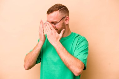 Beyazlı genç bir adam bej arka planda izole edilmiş, elleriyle gözlerini kapatmaktan korkuyor..