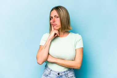 Mavi arka planda izole edilmiş genç beyaz kadın alaycı bir ifadeyle kameraya bakarken mutsuz oluyor..