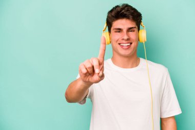 Genç beyaz adam müzik dinliyor. Mavi arka planda izole edilmiş. Bir numarayı parmağıyla gösteriyor..
