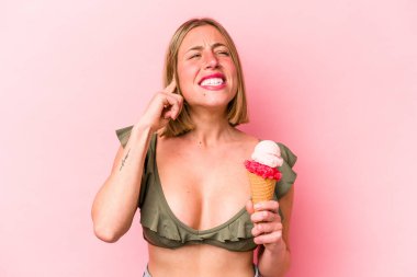 Bikini giyen ve elinde dondurma tutan beyaz bir kadın pembe arka planda izole edilmiş kulakları elleriyle kapatıyor..