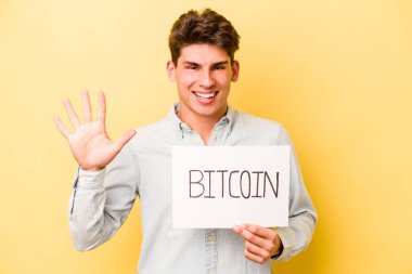 Sarı arka planda bitcoin pankart tutan beyaz bir adam gülümseyerek beş numarayı parmaklarıyla gösteriyor..