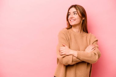 Pembe arka planda izole edilmiş genç beyaz kadın kollarını kavuşturarak gülümsüyor..