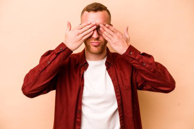 Beyazlı genç bir adam bej arka planda izole edilmiş, elleriyle gözlerini kapatmaktan korkuyor..