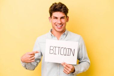 Sarı arka planda izole edilmiş Bitcoin pankartı tutan genç beyaz adam gülümsüyor ve boş alanda bir şey gösteriyor..