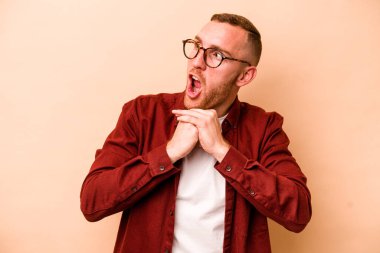 Beyaz tenli genç adam bej arka planda izole edilmiş şans için dua ediyor, şaşırmış ve ağzını açıp önüne bakıyor..