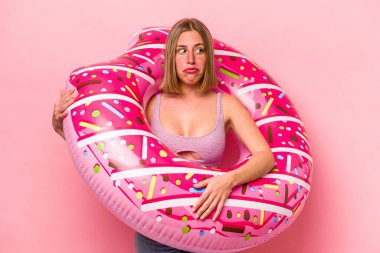 Pembe arka planda izole edilmiş şişme bir yatak tutan genç beyaz kadın kafası karışmış ve kendinden emin değil..