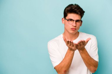 Kafkasyalı genç adam mavi arka planda izole edilmiş dudaklar kıvrılıyor ve hava öpücüğü göndermek için avuç içleri tutuyor..