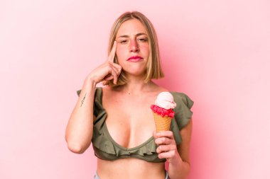 Genç beyaz kadın bikini giyiyor ve pembe arka planda izole edilmiş bir dondurma tutuyor. Şakağı parmağıyla işaret ediyor, düşünüyor ve bir göreve odaklanıyor..