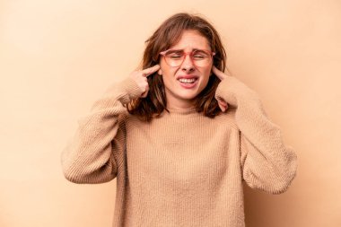 Beyaz tenli genç bir kadın bej arka planda izole edilmiş parmaklarıyla kulaklarını kapatıyor, gürültülü bir ortam yüzünden stresli ve çaresiz..