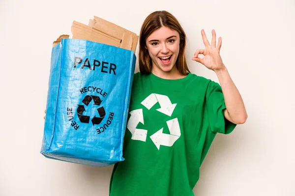 白地に隔離された紙をリサイクルするためにリサイクルバッグを持っている若い女性陽気で自信を持ってOkジェスチャーを示す — ストック写真